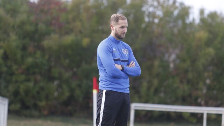 Ein Mann in einem blauen Langarmshirt und schwarzen Hosen mit einem weißen Streifen steht auf einem Feld, beobachtet ein Spiel, mit verschränkten Armen.