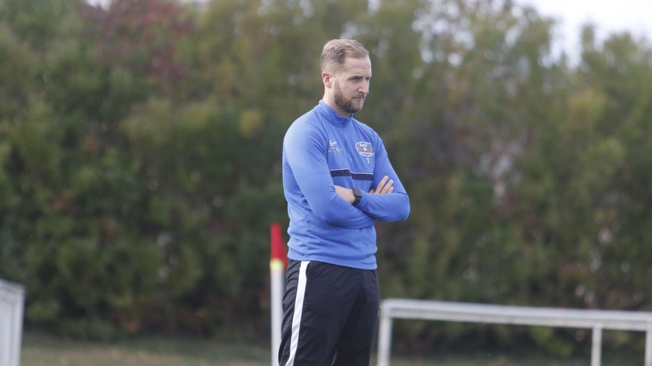 Ein Mann in einem blauen Langarmshirt und schwarzen Hosen mit einem weißen Streifen steht auf einem Feld, beobachtet ein Spiel, mit verschränkten Armen.