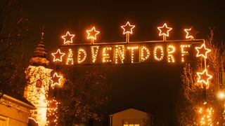 Beleuchtetes Schild mit der Aufschrift 'ADVENTDOR' und fünf Sternen, einer in der Mitte und vier an den Seiten, gelb leuchtend. Darunter ein Haus mit leuchtenden Fenstern.