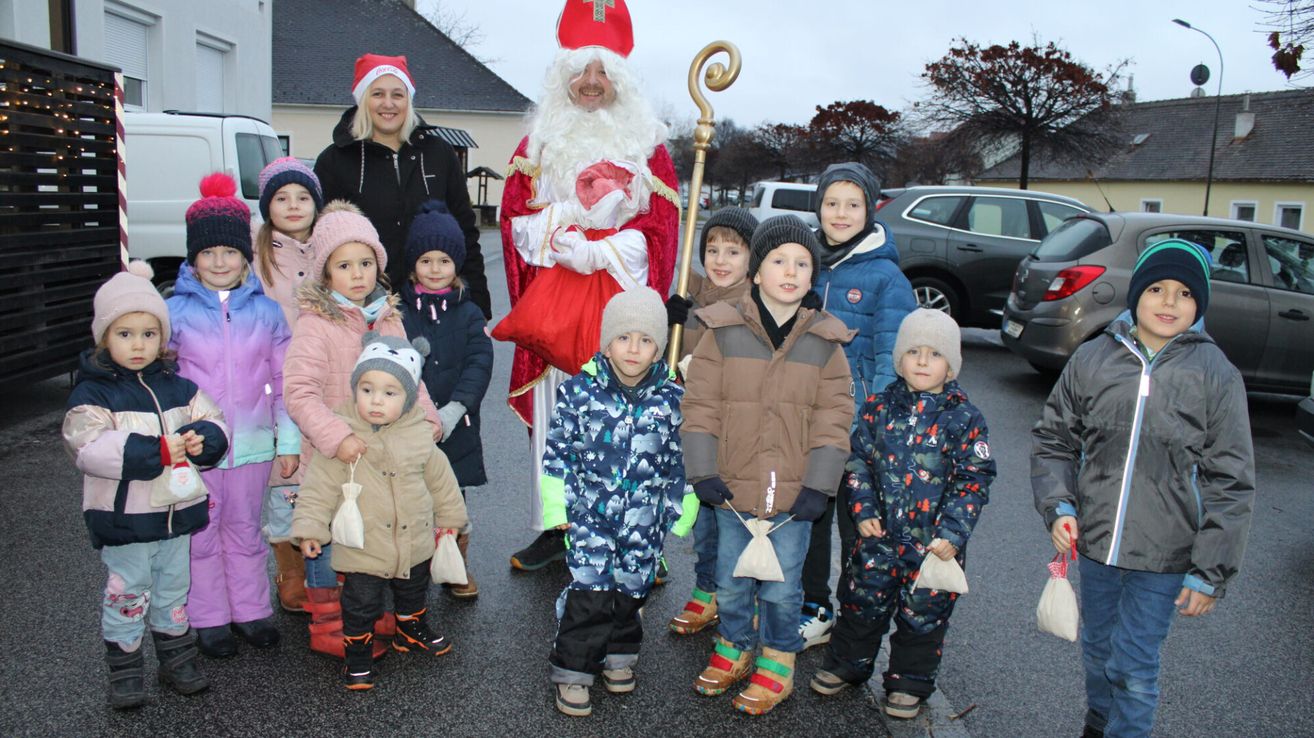 Ein Mann als Weihnachtsmann steht mit Kindern in Winterkleidung. Sie halten kleine Taschen und lächeln, mit Bäumen und einem Haus im Hintergrund.