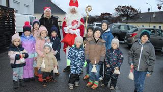Ein Mann als Weihnachtsmann steht mit Kindern in Winterkleidung. Sie halten kleine Taschen und lächeln, mit Bäumen und einem Haus im Hintergrund.