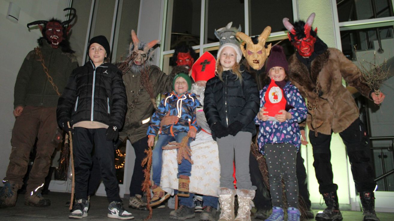 Mehrere Menschen stehen vor einem Gebäude. Ein Junge hält ein Kind auf dem Schoß. Einige von ihnen tragen Masken und halten ein rotes Objekt. Sie tragen alle Winterkleidung. Hinter ihnen sind Lichter.