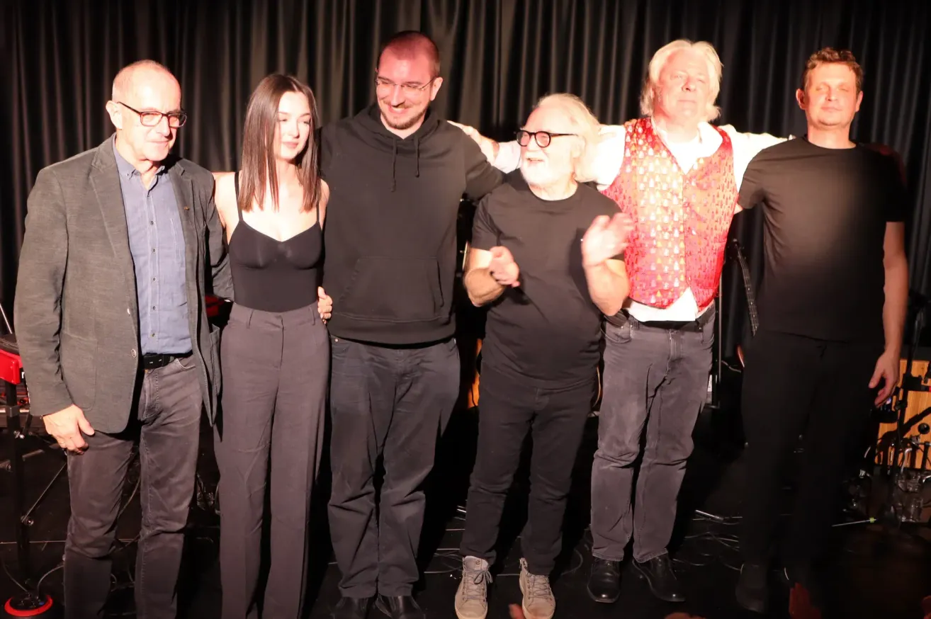 Fünf Personen stehen gemeinsam auf einer Bühne. Der Mann in der Mitte trägt ein rotes Weste und hat seine Arme um zwei andere gelegt. Sie alle tragen Brillen und Turnschuhe.