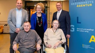 Fünf Personen posieren für ein Foto in einer Innenumgebung. Zwei ältere Personen in Rollstühlen stehen vorne, mit einer Frau in einer blauen Jacke hinter ihnen. Zwei Männer in Anzügen stehen hinter der Frau. Ein blauer Banner mit Text befindet sich auf der rechten Seite.