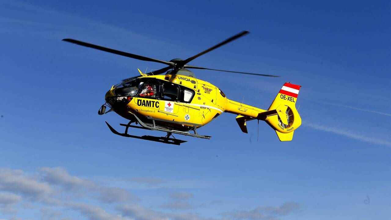 Ein gelber Hubschrauber mit der Aufschrift 'DAMTC' und 'OE-XEL' fliegt in der Luft. Der Hubschrauber hat ein rotes Kreuz auf der Seite.