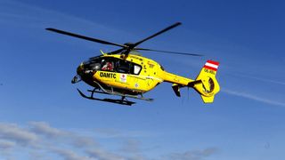 Ein gelber Hubschrauber mit der Aufschrift 'DAMTC' und 'OE-XEL' fliegt in der Luft. Der Hubschrauber hat ein rotes Kreuz auf der Seite.