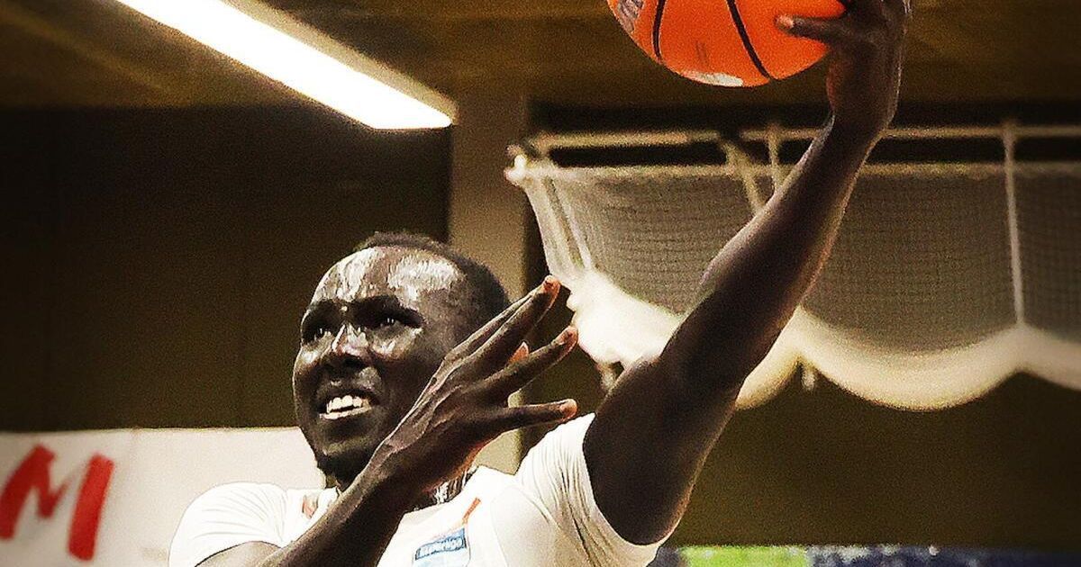 Ein Basketballspieler springt, um den Ball zu werfen. Er trägt ein weißes Trikot und hat einen ernsten Gesichtsausdruck. Im Hintergrund ist der Basketballkorb zu sehen.