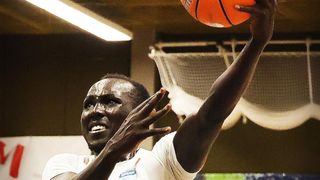 Ein Basketballspieler springt, um den Ball zu werfen. Er trägt ein weißes Trikot und hat einen ernsten Gesichtsausdruck. Im Hintergrund ist der Basketballkorb zu sehen.