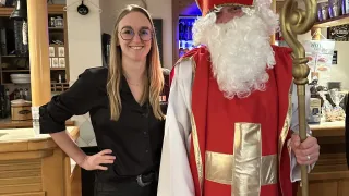 Eine Frau mit Brille und einem schwarzen Oberteil steht neben einem Mann in einem roten und weißen Kostüm mit einem Stab. Sie stehen in einem Restaurant.