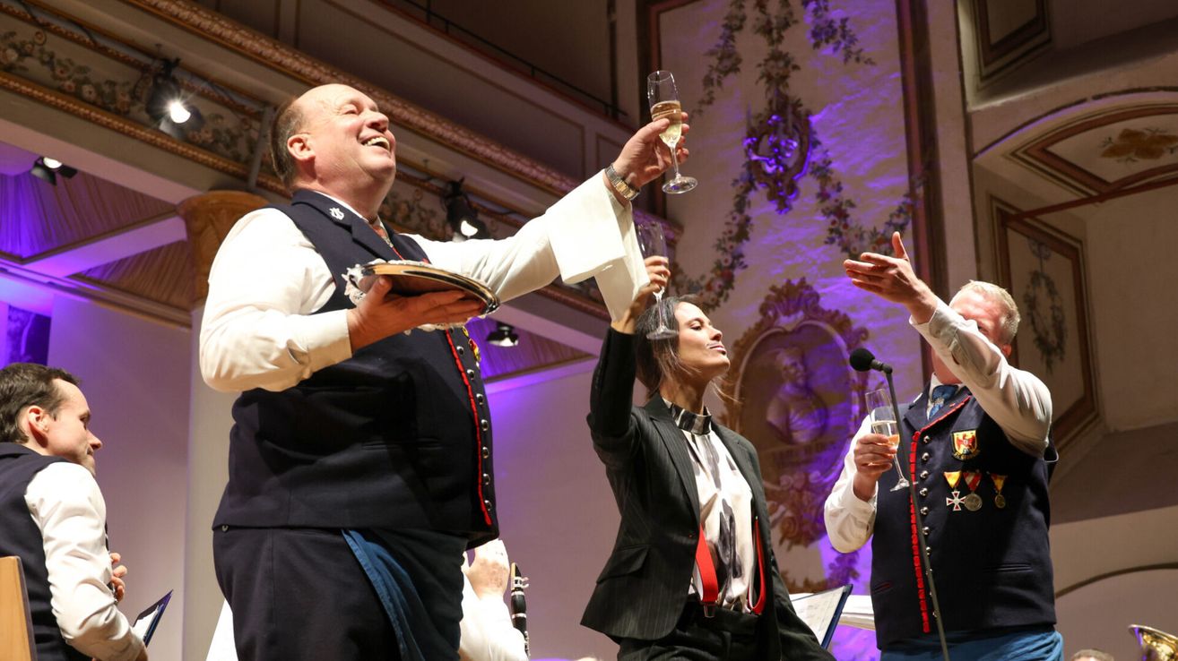 Ein Mann und eine Frau halten Weinglaser zur Feier hoch, mit einem Musiker, der hinter ihnen eine Klarinette spielt.