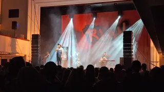 Eine Band tritt auf der Bühne mit roter Beleuchtung und einem großen Hintergrund auf. Ein Sänger steht mit einem Mikrofon in der Mitte, während ein anderer Gitarre spielt. Das Publikum ist im Vordergrund sichtbar.