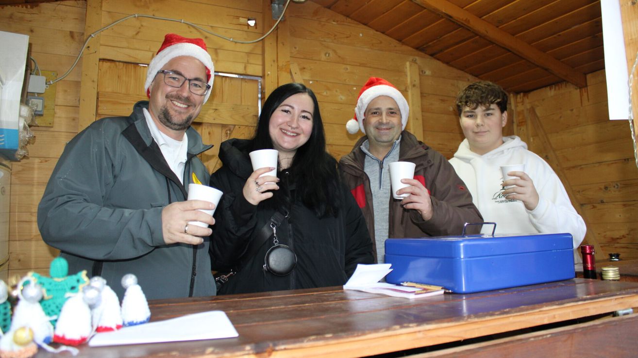 Vier Menschen in Weihnachtsmützen stehen hinter einem Holztisch und halten Tassen. Die Frau in der Mitte lächelt. Auf dem Tisch liegen Papiere, eine Schachtel und ein Stift. Die Wand hinter ihnen ist aus Holz.