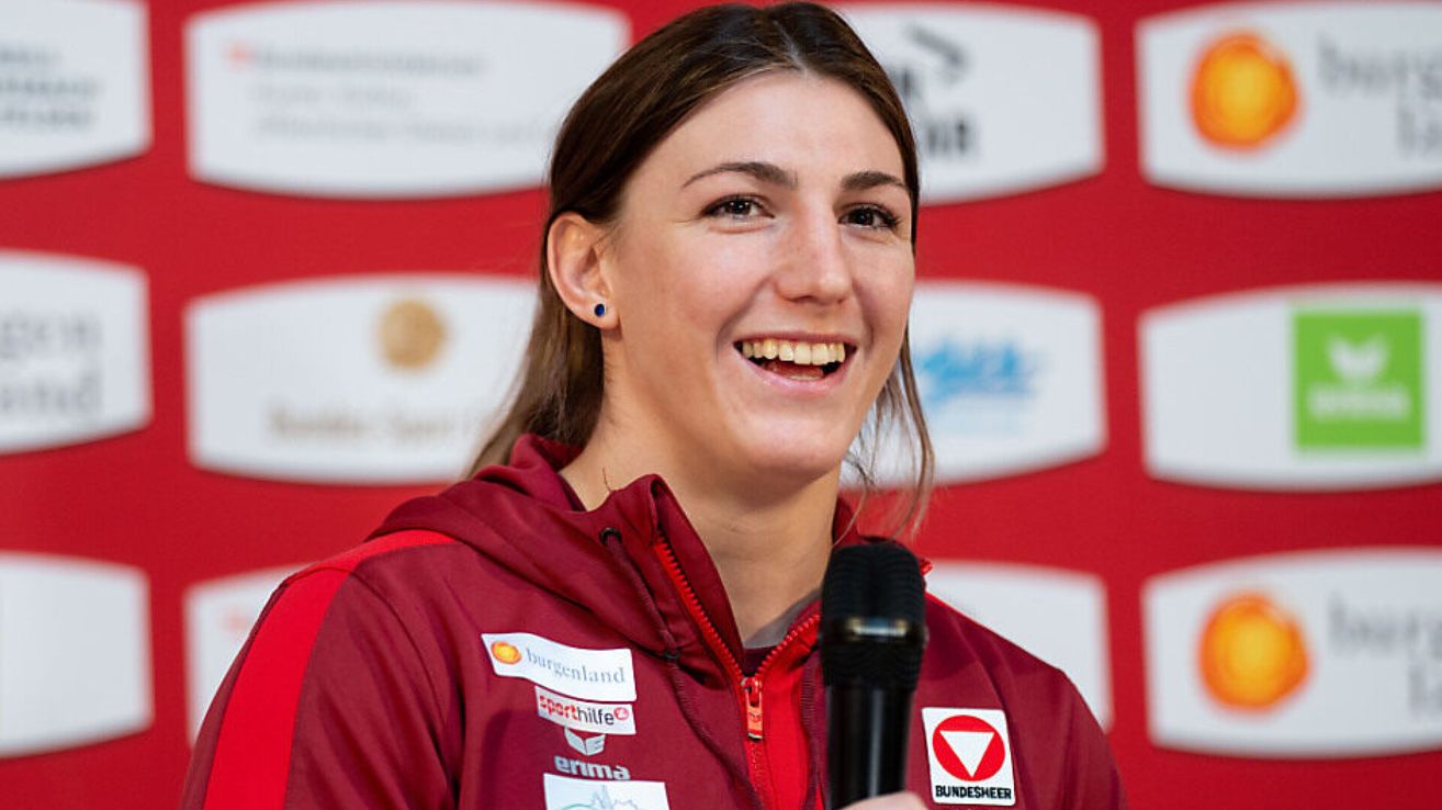 Eine Frau mit braunen Haaren, die breit lächelt, spricht in ein Mikrofon. Sie trägt eine rote Jacke mit mehreren Logos und Aufnähern.