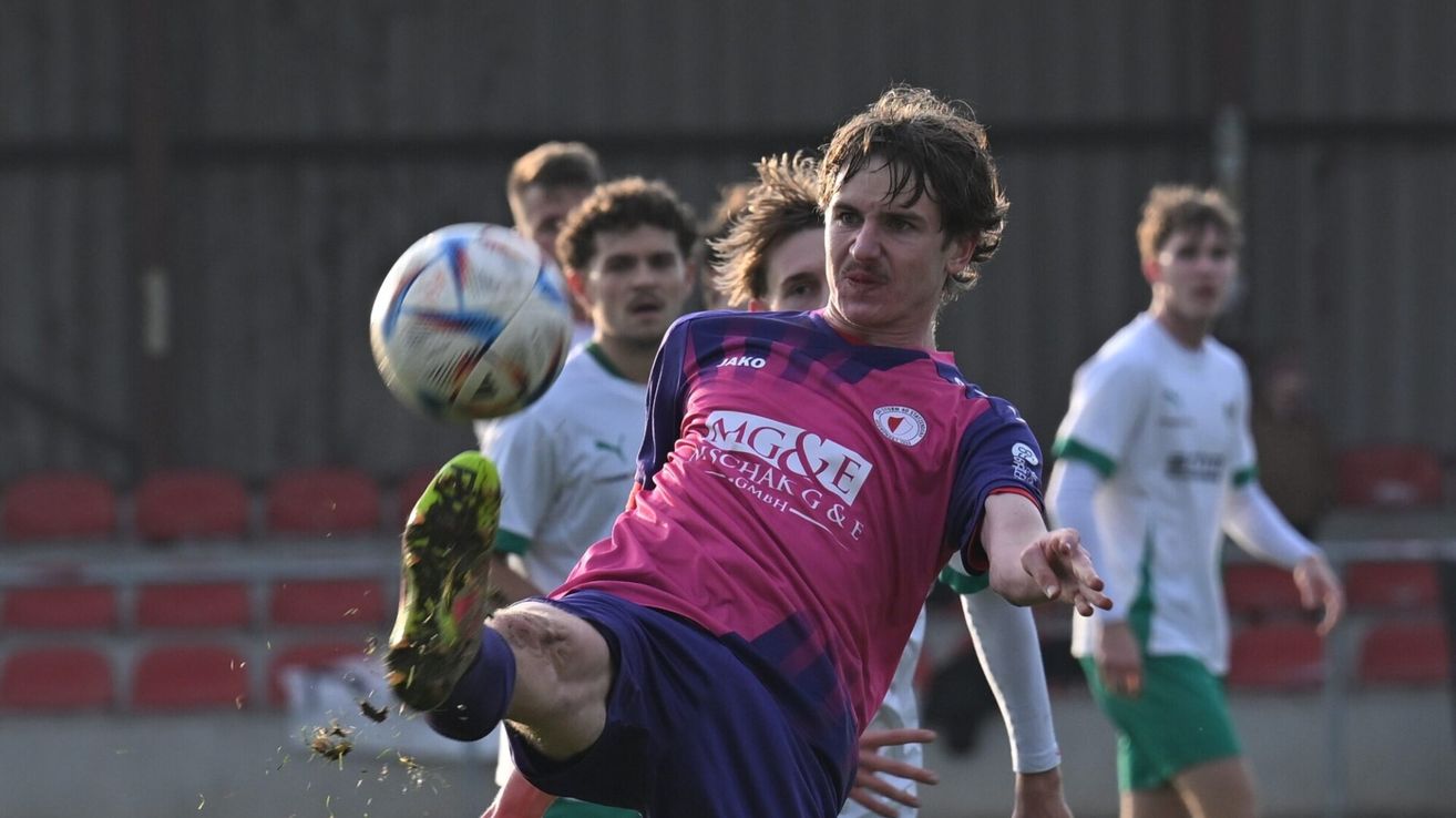 Ein Fußballspieler tritt einen Ball in der Luft, während seine Teamkollegen zusehen. Der Spieler trägt ein pinkes und blaues Trikot. Der Hintergrund zeigt ein verschwommenes Stadion.
