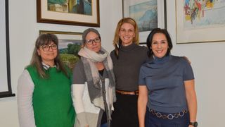 Vier Frauen stehen vor einer Wand mit Gemälden. Sie lächeln. Eine trägt eine Schürze, eine andere hat eine Kopfbinde. Die Wand hat zwei gerahmte Bilder.