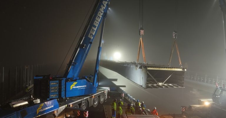 Ein Kran hebt bei Nacht eine große Betonstruktur. Arbeiter in Sicherheitsausrüstung stehen in der Nähe. Der Kran ist blau mit 'FELBERMAYR' darauf geschrieben.