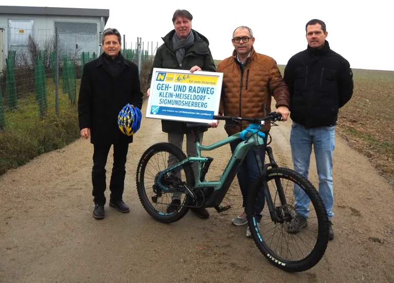 Vier Männer stehen neben einem Fahrrad auf einem Feldweg. Der Mann in der Mitte hält ein Schild, auf dem Geh- und Radweg Klein-Meiseldorf - Sigmundsherberg steht.