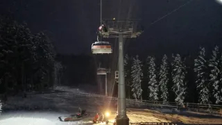 Eine Skiliftstation ist bei Nacht zu sehen, mit zwei Personen in roten Jacken, die in der Nähe der Basis stehen. Ein Schneemobil ist geparkt. Das Gebiet ist von schneebedeckten Bäumen und einem Zaun umgeben.