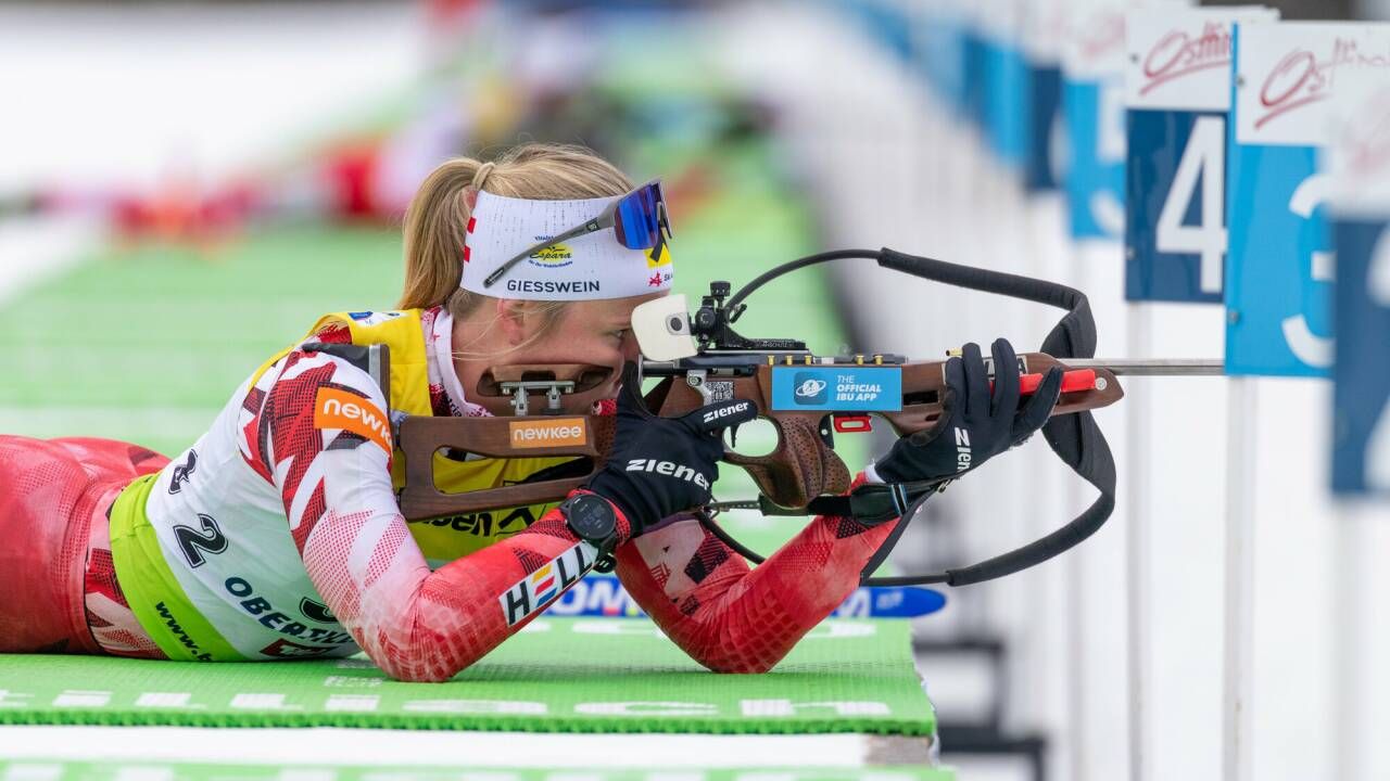 Eine weibliche Biathletin liegt auf dem Boden und zielt mit einem Gewehr mit einem blauen Aufkleber, der 'The Official IBU App' beschriftet ist. Sie trägt eine weiße Kopfband, eine Schutzbrille und gelbe und rote Sportbekleidung.