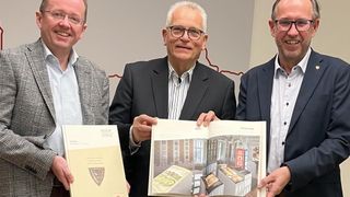 Drei Männer in Anzügen halten Bücher und lächeln, eines der Bücher zeigt eine Zeichnung eines Gebäudes.