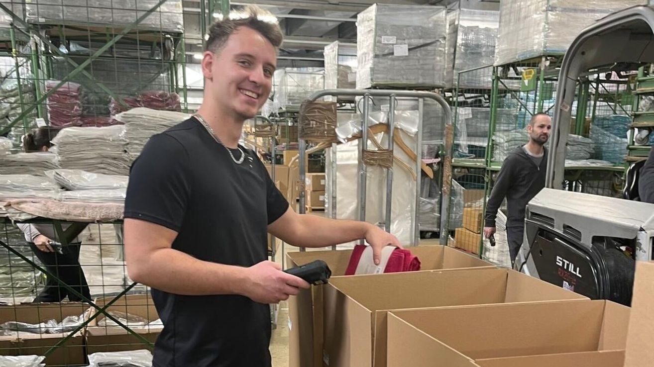 Ein Mann in einem Lagerraum hält einen Scanner und betrachtet Kisten. Er lächelt und scheint seine Arbeit zu genießen.