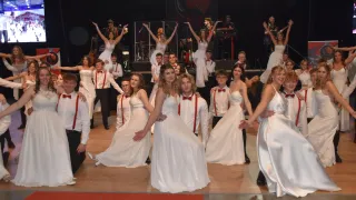 Eine Gruppe von Menschen tritt auf der Bühne auf, gekleidet in weiße Kleider und rote Fliegen. Sie scheinen in einer koordinierten Weise zu tanzen. Einige von ihnen spielen Musikinstrumente, während andere hinter ihnen stehen.