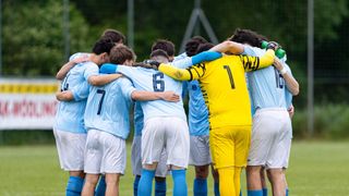 Eine Fußballmannschaft in blauen und weißen Trikots ist auf einem Feld versammelt. Der Spieler in Gelb trägt die Nummer 1.