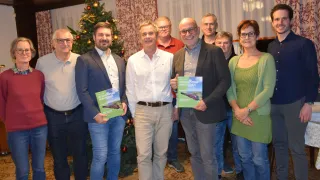 Eine Gruppe von Menschen steht zusammen in einem Raum. Einige halten Bücher, während andere für die Kamera lächeln. Ein Weihnachtsbaum ist im Hintergrund.