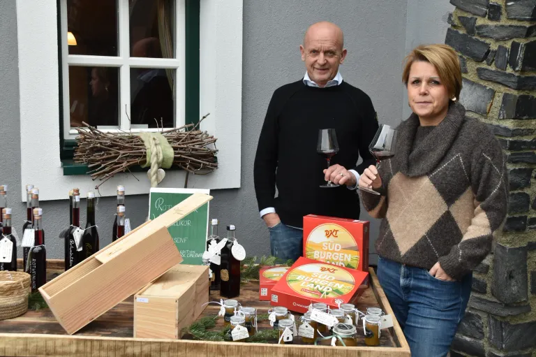 Ein Mann und eine Frau stehen vor einem Tisch mit Weingläsern, Kisten und Gläsern. Der Mann hält eine Flasche und die Frau lächelt. Ein Kranz hängt an der Wand hinter ihnen.