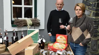 Ein Mann und eine Frau stehen vor einem Tisch mit Weingläsern, Kisten und Gläsern. Der Mann hält eine Flasche und die Frau lächelt. Ein Kranz hängt an der Wand hinter ihnen.
