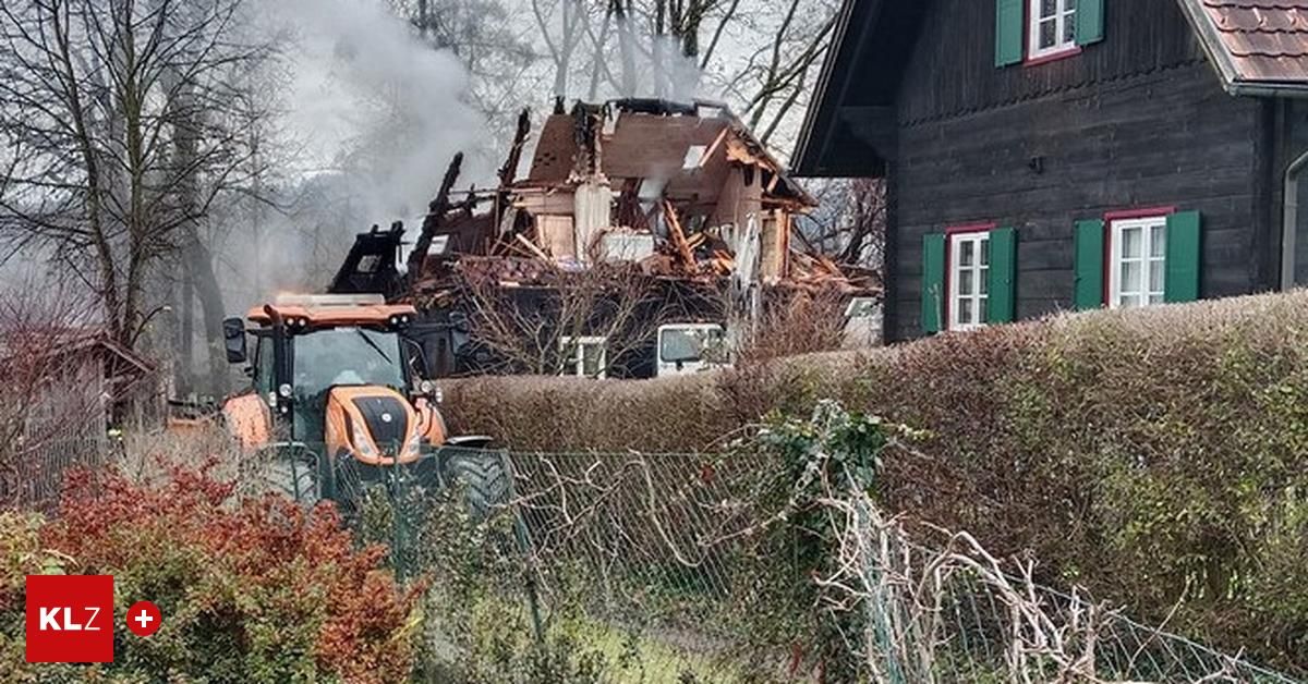 Ein Traktor steht vor einem teilweise zerstörten Haus, aus dem Rauch aufsteigt. Das Haus ist von Bäumen und Büschen umgeben.