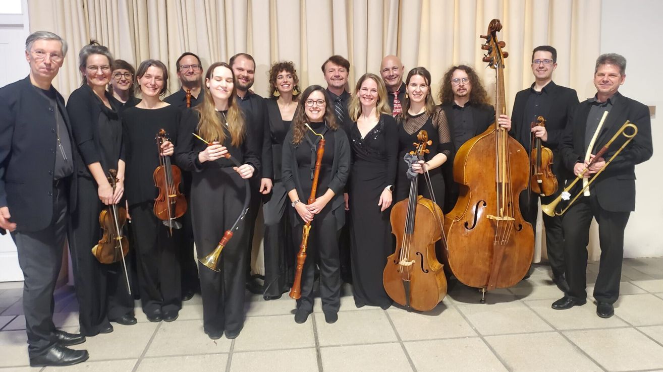 Eine Gruppe von Musikern, alle in Schwarz gekleidet, posiert für ein Foto. Sie halten verschiedene Musikinstrumente, darunter Geigen, Violoncelli und Flöten. Der Hintergrund ist formell, mit einem Vorhang.