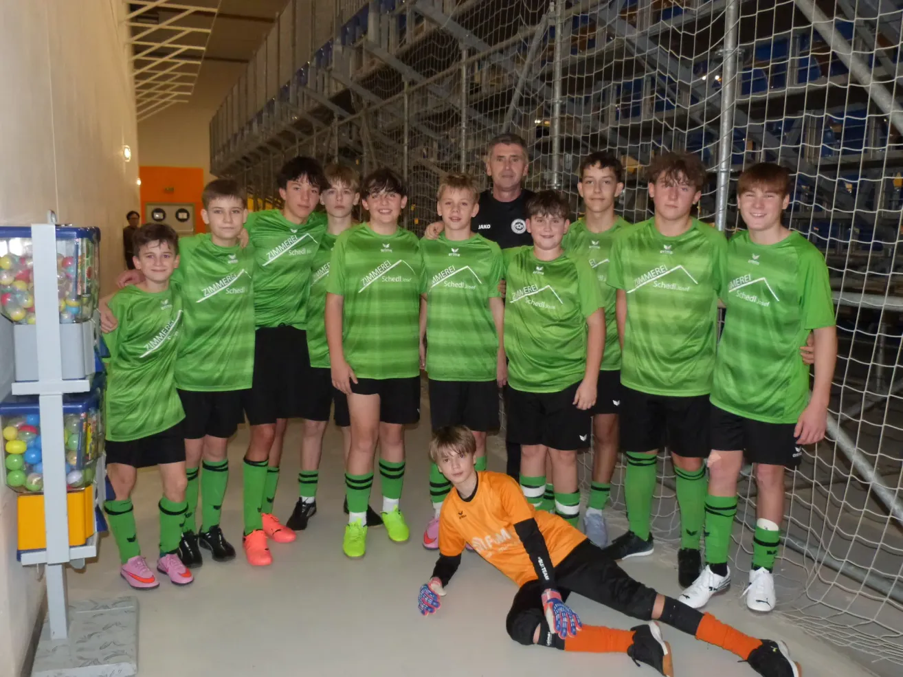 Eine Gruppe junger Fußballer in grünen Trikots und schwarzen Shorts steht vor einem Tor mit einem Mann in einem schwarzen Shirt dahinter. Ein Junge in einem orangefarbenen Shirt kniet vor dem Tor.