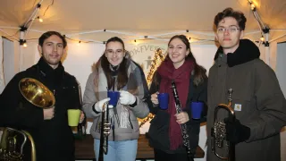 Vier Personen stehen in einem Raum mit Musikinstrumenten und blauen Bechern. Dahinter befindet sich ein Weihnachtsbaum.