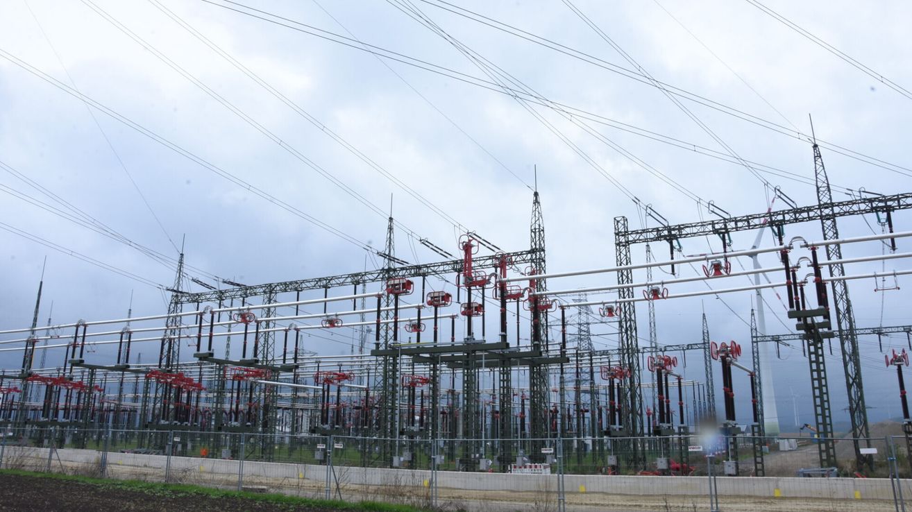 Ein ausgedehntes Kraftwerk unter einem bewölkten Himmel mit mehreren hohen Türmen mit roten und weißen Komponenten, die durch zahlreiche Drähte miteinander verbunden sind.