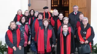 Eine Gruppe älterer Frauen und Männer, gekleidet in rote und schwarze Mäntel, posiert für ein Foto vor einer Holztür und lächelt in die Kamera.