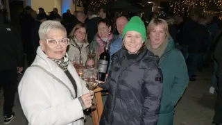 Eine Gruppe von Menschen in Winterkleidung lächelt und posiert für ein Foto. Sie halten Weingläser und Flaschen und feiern möglicherweise ein Ereignis. Die Frau links trägt eine Brille. Die Frau in der Mitte trägt eine grüne Mütze und hält eine Flasche. Hinter ihnen stehen weitere Menschen, von denen einige verschwommen sind.
