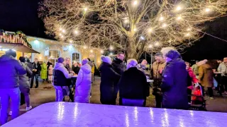 In der Nacht versammeln sich mehrere Personen um einen Tisch unter einem Baum mit dekorativen Lichtern und unterhalten sich. Der Bereich wird von Straßenlaternen und dem Gebäude dahinter beleuchtet.