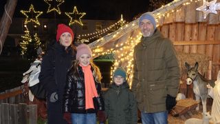 Eine vierköpfige Familie steht vor einer dekorierten Holzstruktur mit Lichtern. Die Erwachsenen tragen Winterkleidung und Mützen, während die Kinder Schals und Handschuhe haben. Sie scheinen für ein Foto zu posieren.