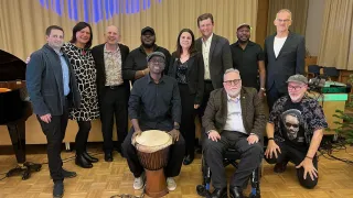 Eine Gruppe von Menschen steht und sitzt für ein Foto in einem Konferenzraum zusammen. Ein Mann spielt eine Trommel.