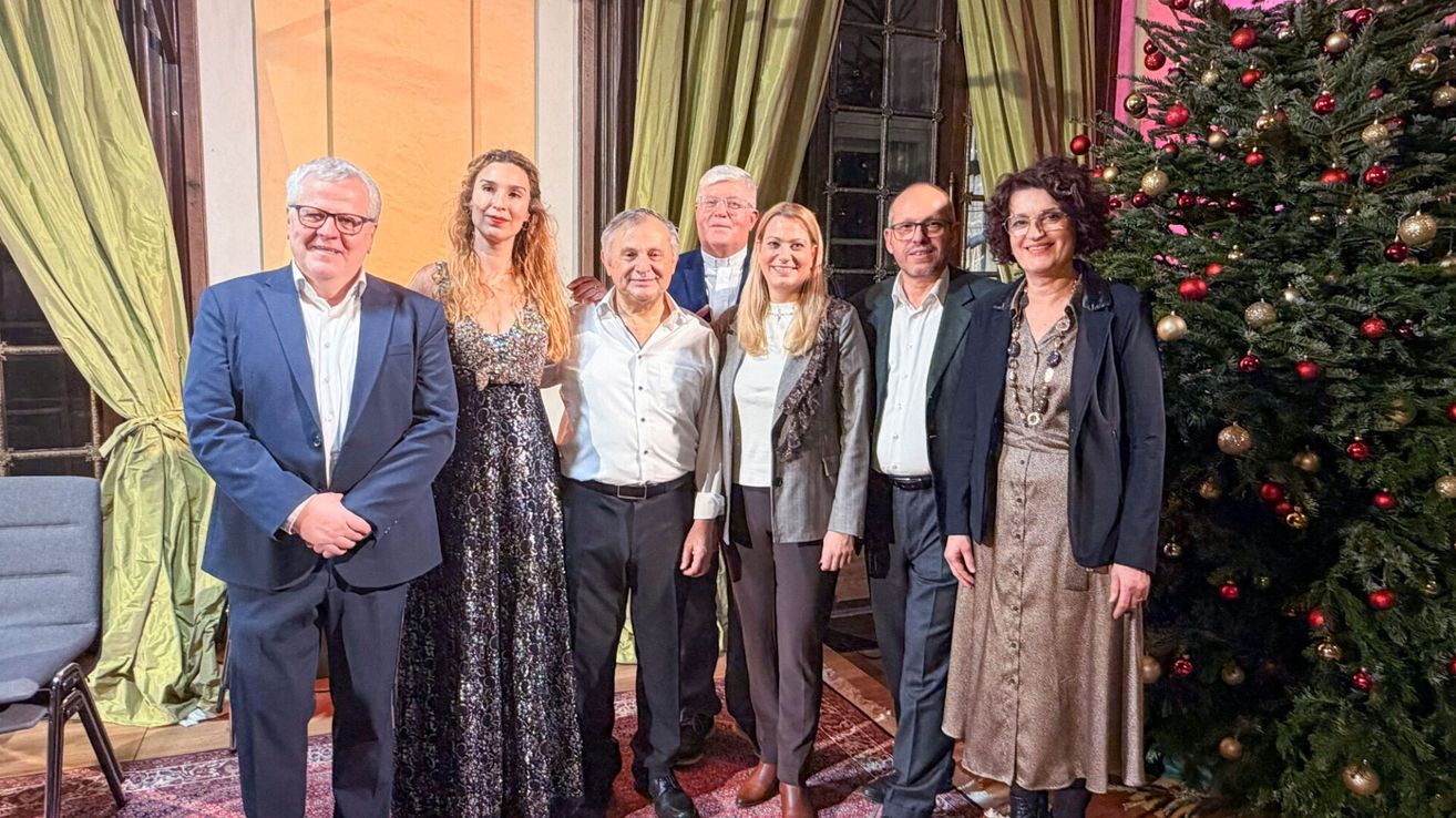 Eine Gruppe von Menschen, wahrscheinlich Familie, posiert für ein Foto vor einem geschmückten Weihnachtsbaum. Sie sind formell gekleidet, einige tragen Brillen. Hinter ihnen befindet sich ein Fenster mit Vorhängen.