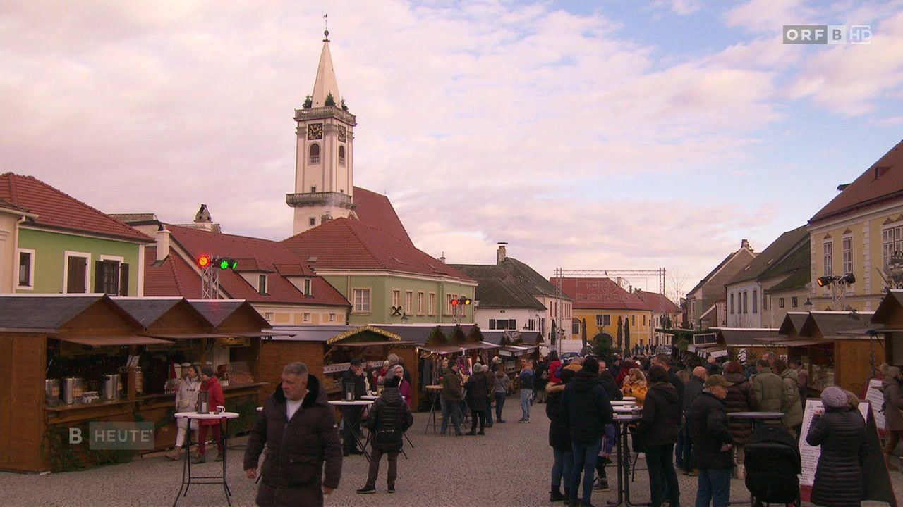 Ein geschäftiger Stadtplatz voller Menschen. Bunte Stände säumen die Straße, und ein hoher Turm mit einer Uhr ist im Hintergrund zu sehen.