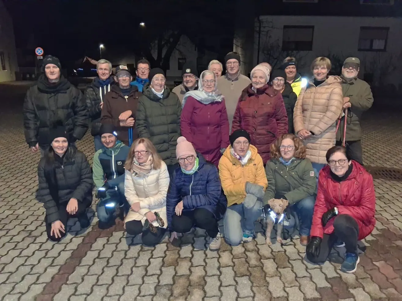 Eine Gruppe von Menschen in Winterkleidung posiert nachts draußen für ein Foto. Sie lächeln und einige haben Winteraccessoires. Ein Hund sitzt vor ihnen.