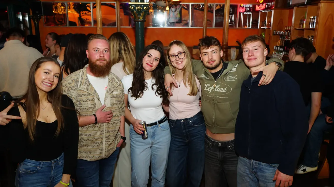 Eine Gruppe von fünf Freunden steht zusammen in einer Bar und posiert für ein Foto. Ein Mann trägt eine grüne Jacke mit Logo. Zwei Frauen tragen eine Brille und eine hält eine Dose. Sie lächeln und scheinen den Moment zu genießen.