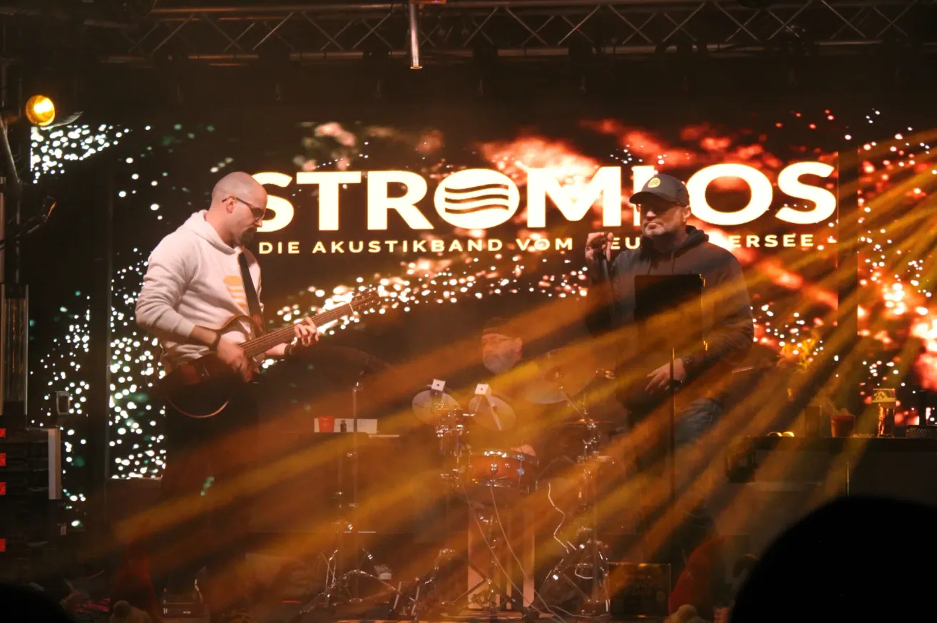 Eine Band auf der Bühne mit einem Gitarristen, einem Schlagzeuger und einem Sänger, alles unter einem großen Banner mit dem Text 'Stromio'.