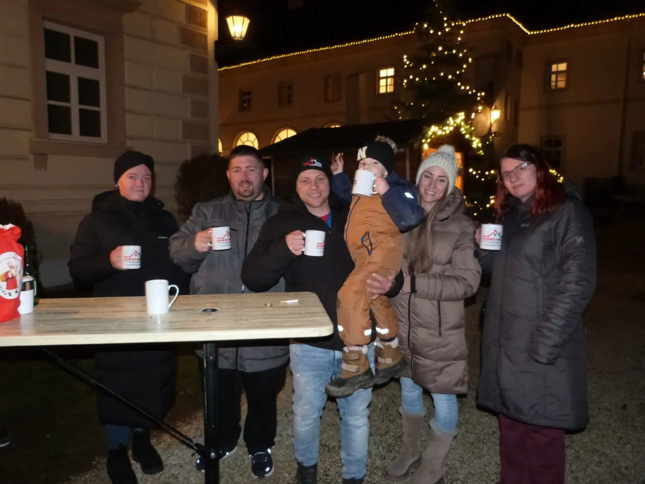A family of six, dressed in winter clothing, stands in front of a lit building, holding mugs, smiling for a photo.