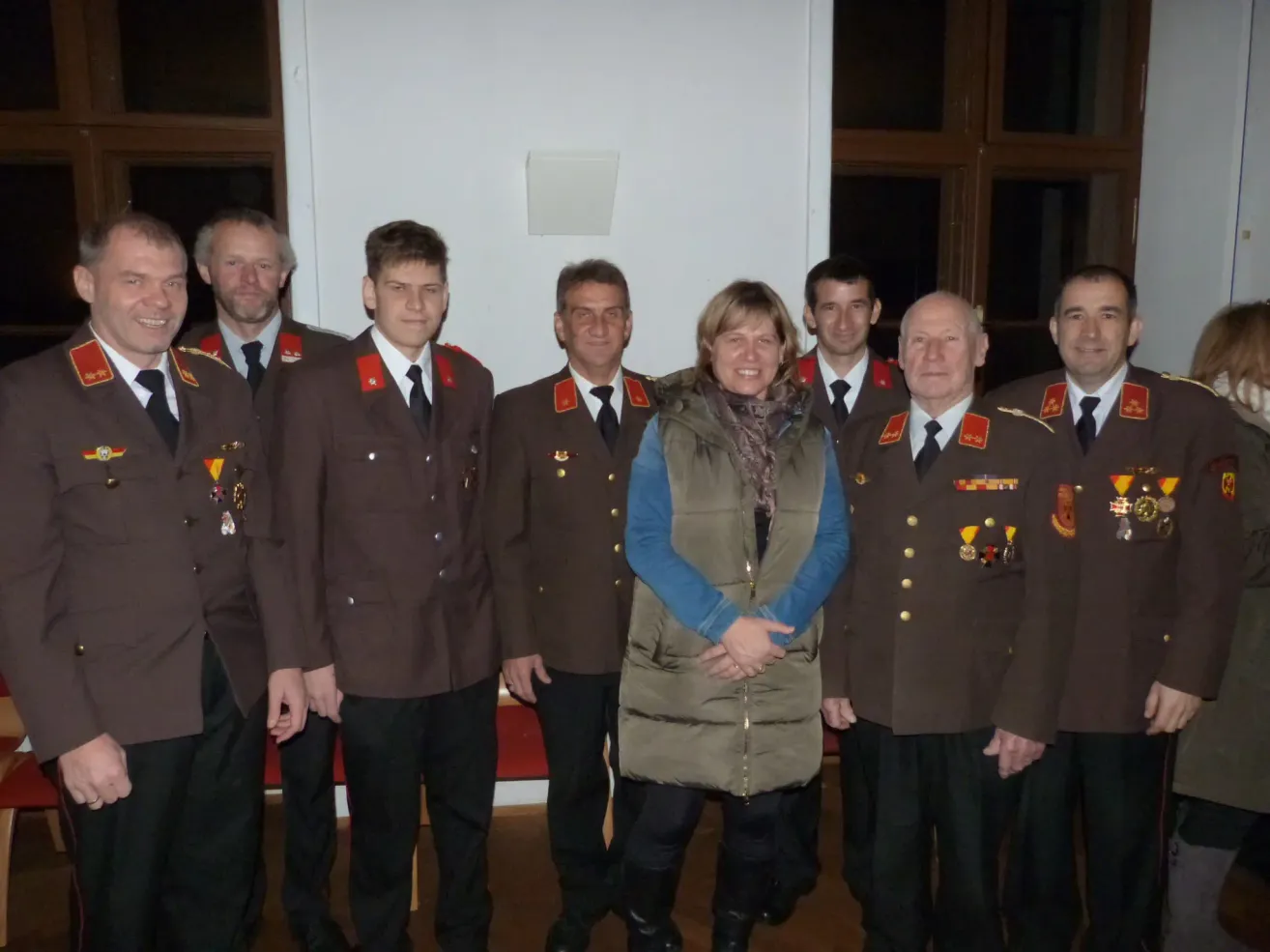 A group of people in uniform and medals stand together, including a woman in a jacket, in front of a white wall.