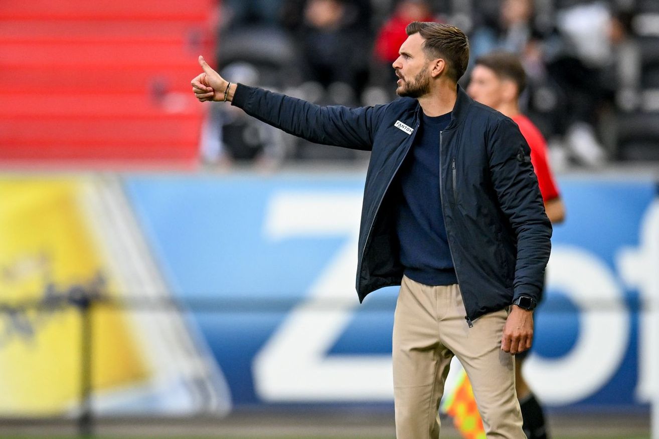 Ein Fußballtrainer in einer blauen Jacke gestikuliert mit der rechten Hand auf dem Spielfeld. Im Hintergrund sehen verwaschene Zuschauer auf roten und blauen Tribünen.