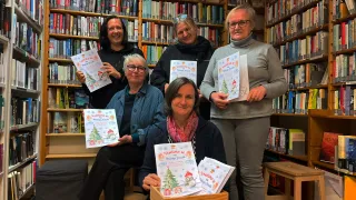 Fünf Frauen in einer Bibliothek posieren mit Weihnachtsbüchern, lächeln und halten die Bücher. Im Hintergrund sind mit Büchern gefüllte Regale sichtbar.