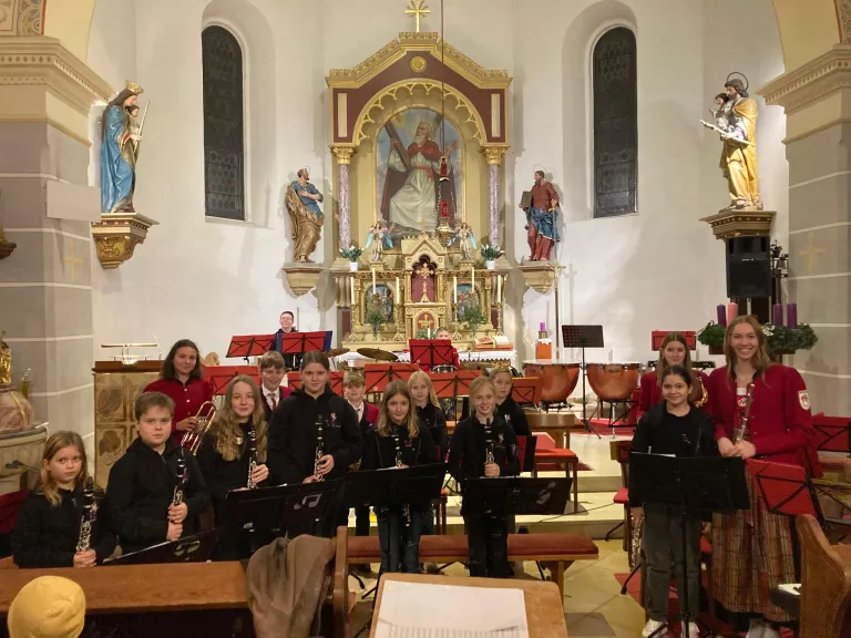 A group of children stand in a church, holding musical instruments, likely clarinets, in front of a decorated altar and statues.